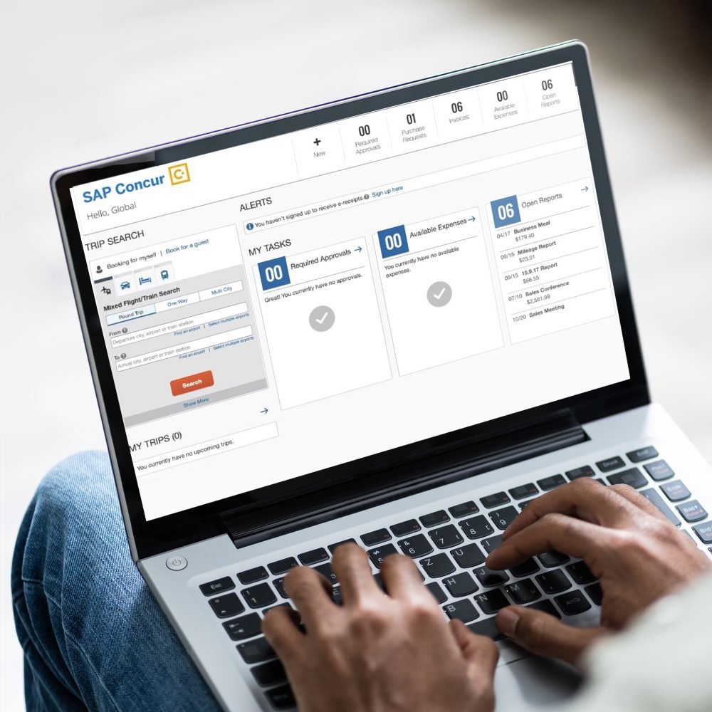 laptop sitting on legs of person wearing jeans; black hands resting on keyboard; SAP Concur screen open on laptop screen